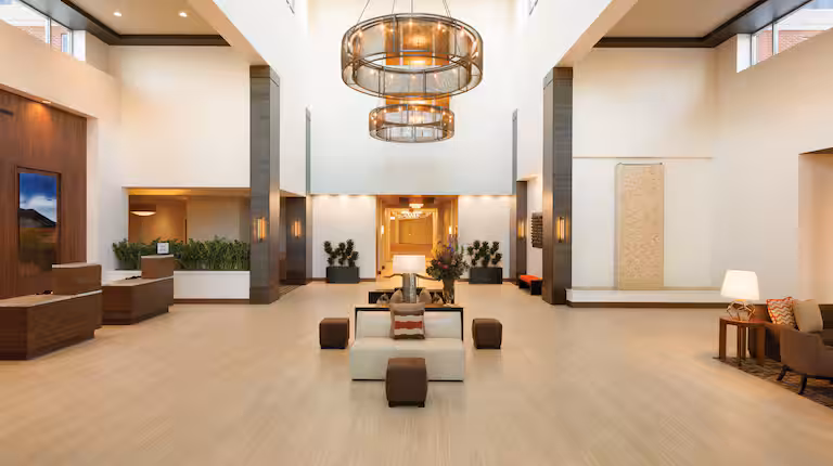 Embassy_Suites_OKC_Atrium A hotel lobby with white walls and tan hardwood floor. a white couch in front of a decorative table sits below a two-tiered circular light fixture centered in frame